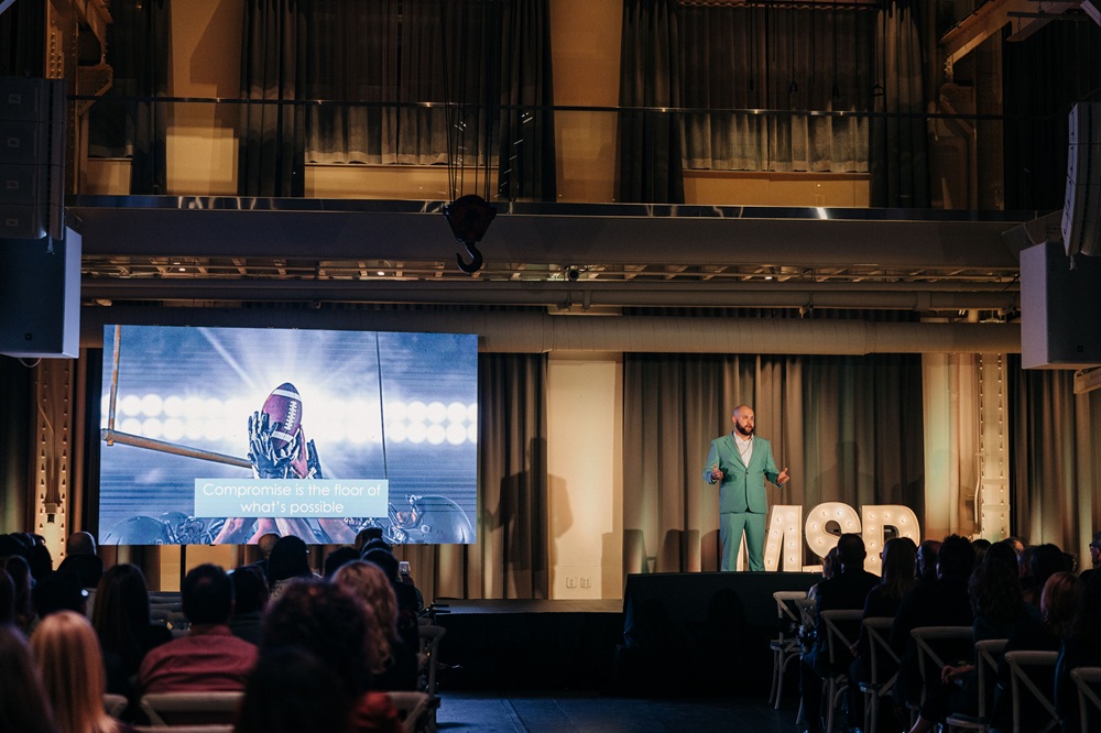 Steve Reuter speaking on stage at DisruptHR, the slide behind him reading 'Compromise is the floor of what's possible'